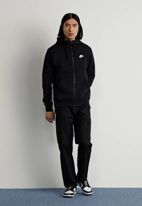 Felpa nera con zip e piccolo logo Nike bianco, abbinata a pantaloni cargo neri e sneakers bianche e nere. Tessuto dalla texture liscia.