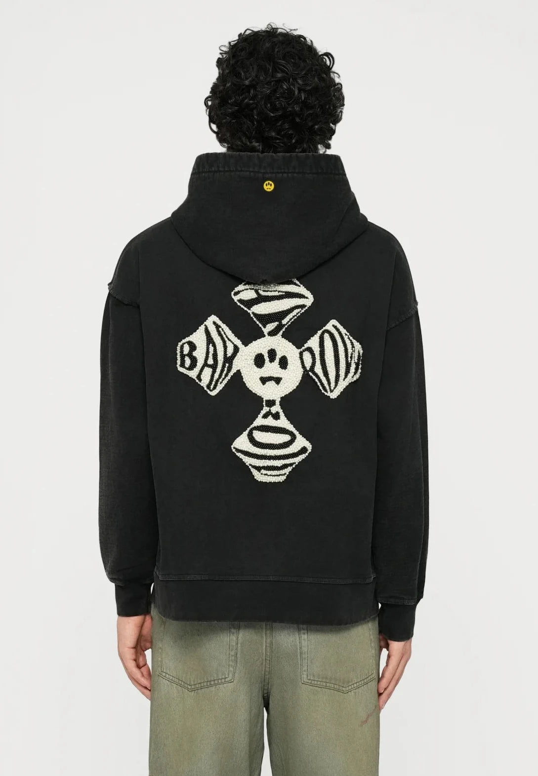 Black hoodie with back graphic design featuring a smiling face and circular patterns. Worn with loose-fitting light green jeans and white sneakers.