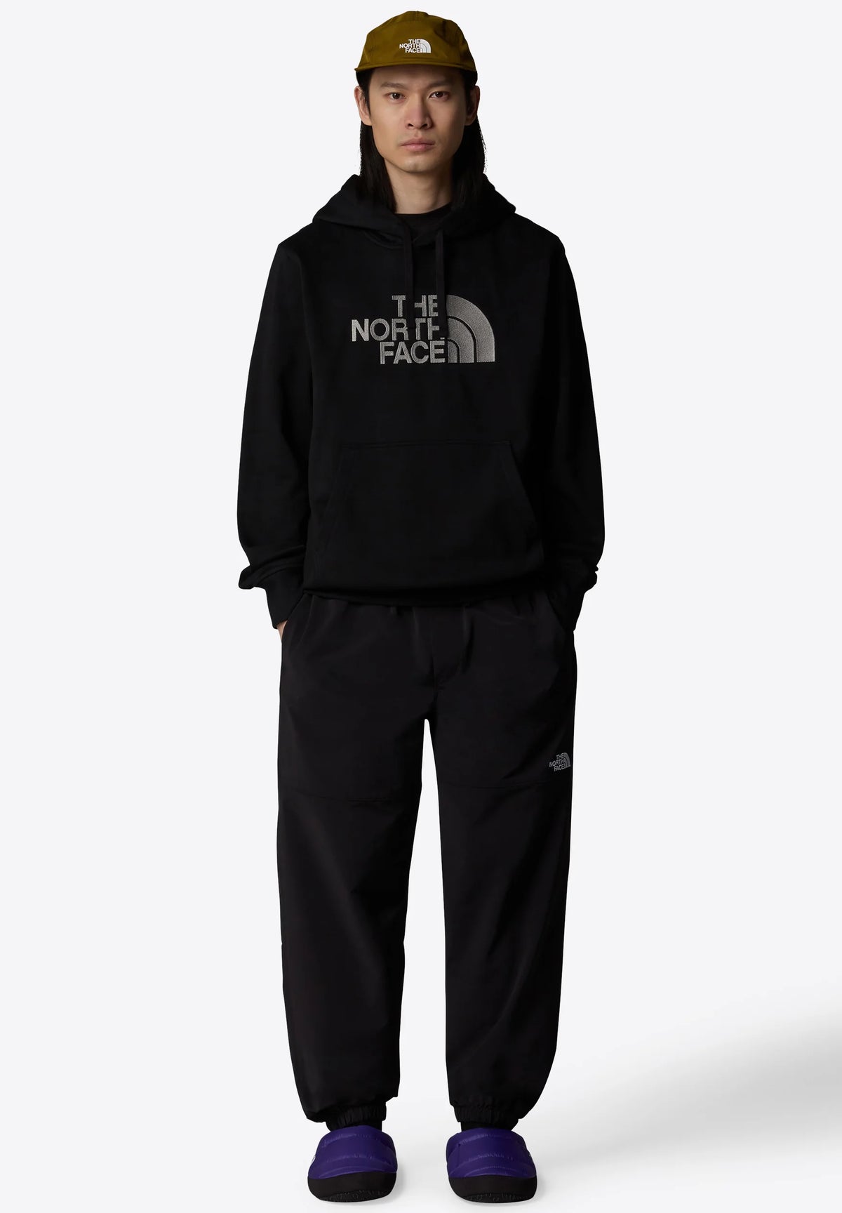 Hoodie nero con un logo grigio prominente, pantaloni neri e pantofole viola. Accessorio: cappellino verde oliva con logo. Vestibilità rilassata, stile casual.