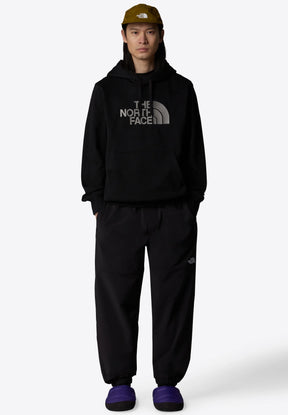 Hoodie nero con un logo grigio prominente, pantaloni neri e pantofole viola. Accessorio: cappellino verde oliva con logo. Vestibilità rilassata, stile casual.