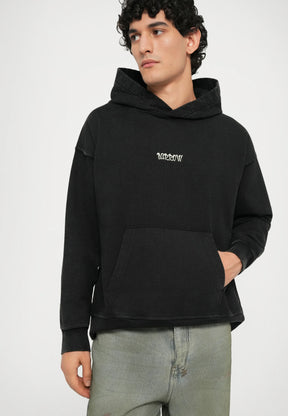 Black hooded sweatshirt with a front pocket, featuring subtle frayed edges and light embroidery on the chest. Worn with light green jeans.