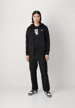 Felpa nera con zip e tasche frontali, abbinata a una maglietta nera con logo bianco; pantaloni cargo neri e sneakers nere/bianche.